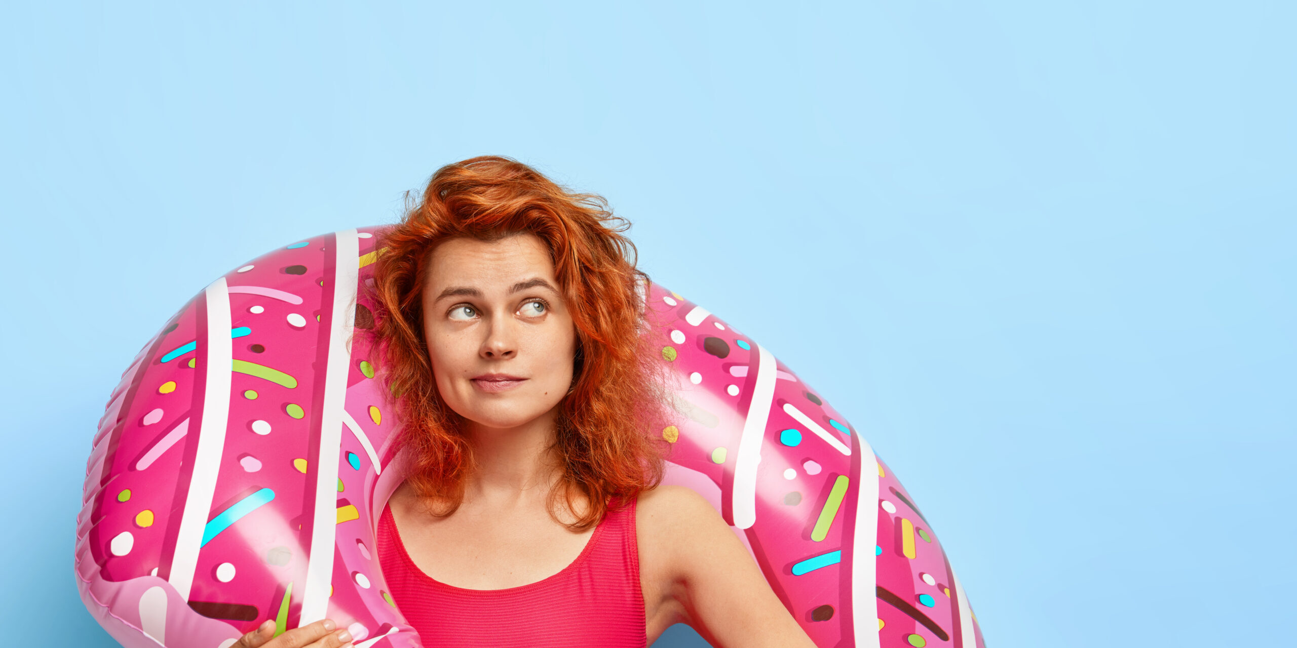 Vertical shot of lovely red haired woman with dimples on cheeks, focused aside, plans future vacation, wears red bikini, holds colorful inflated ring, ready for good rest, isolated on blue background